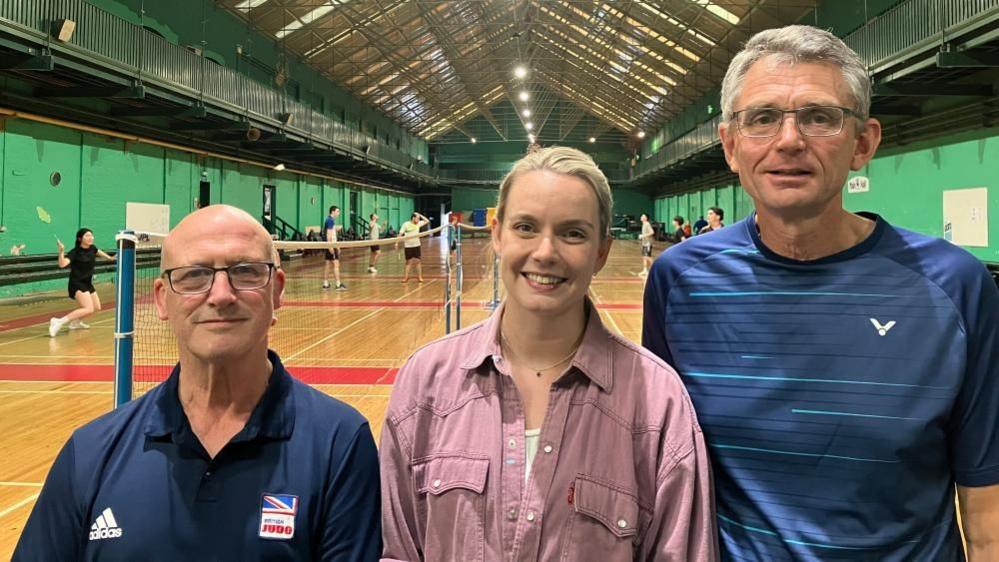 Dermott Heslop, Heather Robertson and Mike Poulter stand side-by-side in front of badminton courts, where players can be seen having a game. Mr Heslop wears glasses and a Navy sports top with a British Judo logo on the chest, Ms Robertson wears a pink shirt and Mr Poulter has a navy blue sports top on, and glasses.