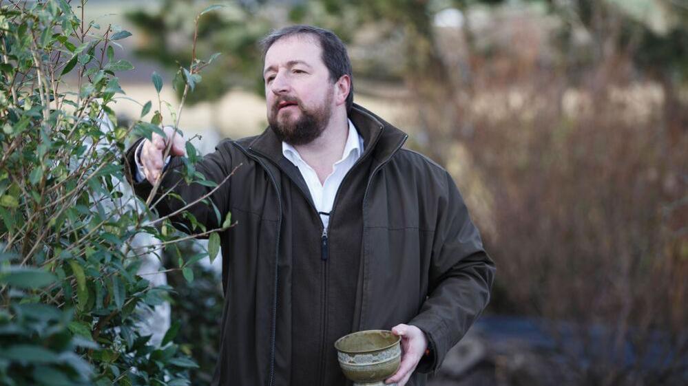 Thomas Robinson collects tea from a field. He is dressed in a white shirt, brown fleece and a brown wax jacket