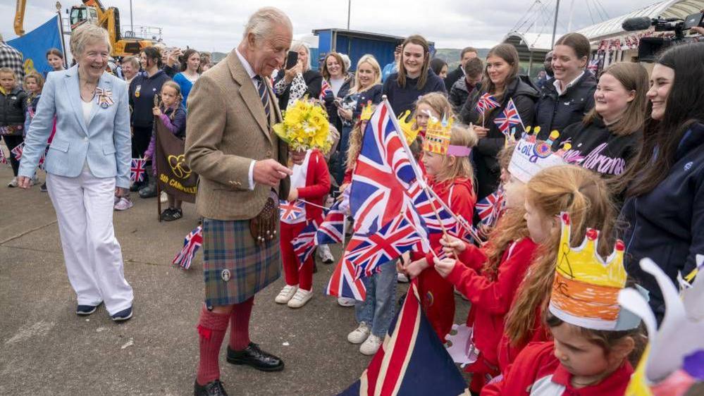 King Charles wearing a kilt being greeting with a crowd of young children who are wearing handmade crowns and waving union jacks