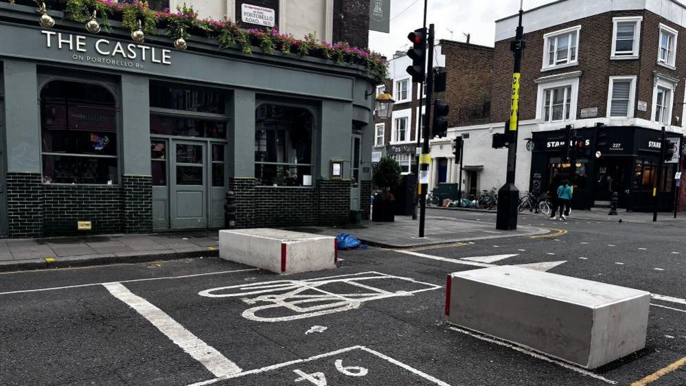 A street junction from one side of the road. The road has white markings, displaying a traffic light crossing, a bicycle stopping zone, and parking space, as well as double yellow lines around the edges of the pavement. There are two grey concrete blocks blocking the road. A pavement made of grey paving slabs runs alongside the road. Across the street, there is a blue-grey building with 'The Castle' written over it. A turning to the left in the background has another street, with brown brick buildings lining it.