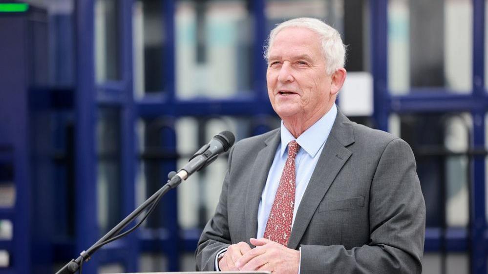 Glen Sanderson speaking into a microphone. He has white, short hair and is wearing a grey suit, light blue shirt and red patterned tie. His hands are crossed in front of him.
