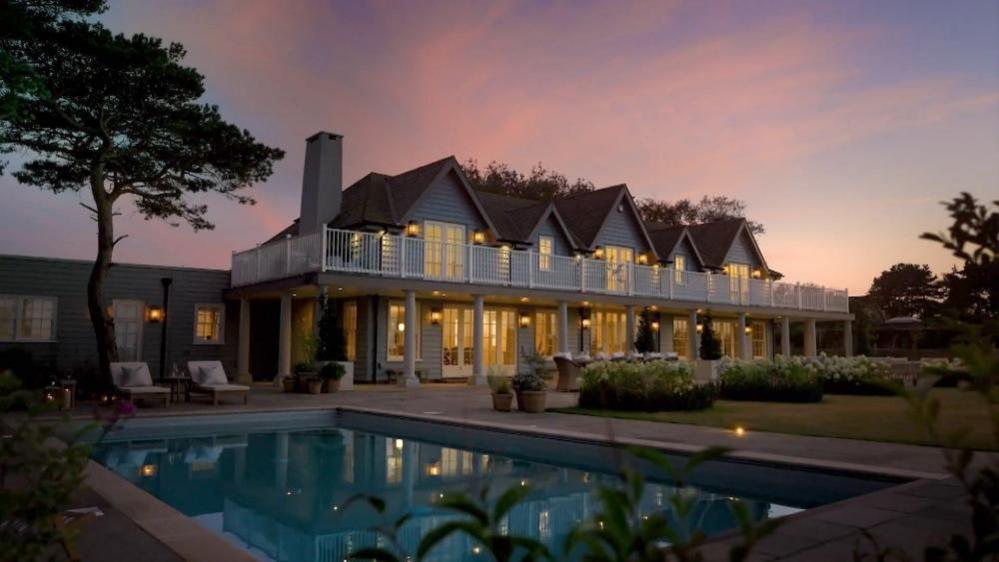 A nighttime image of Larkfields, used in the Omaze marketing campaign. The image is set over the swimming pool, looking to the rear of the house, with an evening sunset sky above.
