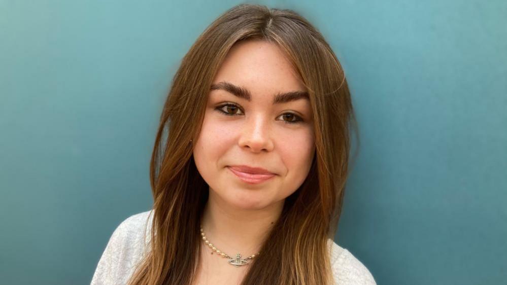 Amie, standing in front of a green wall, looking straight at the camera. She has long dark hair, with a middle parting, and has on a necklace and a grey top.