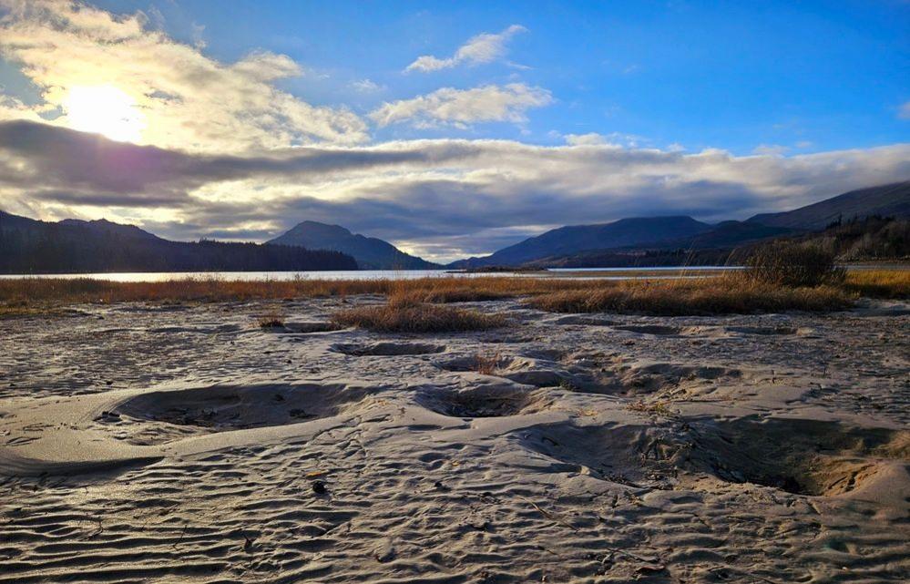 A low down angle of a beach with little imprints in the sand and brown sprouts of plants scattered across the foreground. The view looks up towards hills in the background. It's a cloudy day with patches of blue sky.