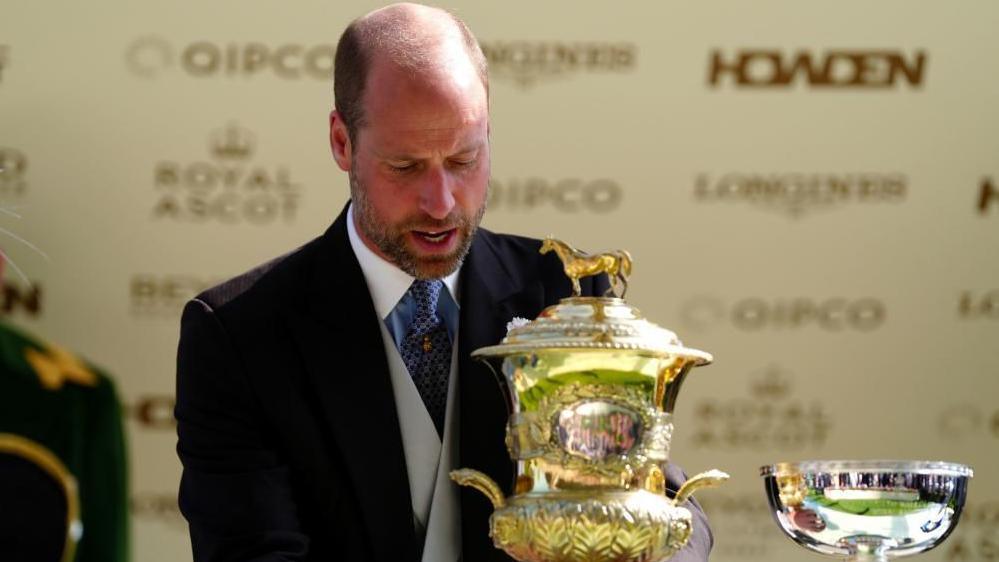 Prince William holds an ornate gold trophy. He is wearing a black dinner jacket, with a light grey waistcoast underneath, white and blue shirt with a blue tie.
