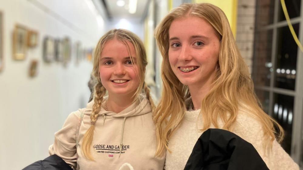 Two young women with blonde hair smile at the camera, standing in a corridor.