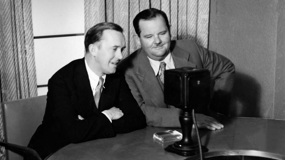 Stan Laurel and Oliver Hardy are sitting at a table with what appears to be a microphone in front of them. They are both smiling and looking at the microphone. There are opened diagonal striped curtains on the window behind them. Stan Laurel has has arms folded on the table, while Oliver Hardy has his left arm on his waist and his other hand flat on the table top. Both are wearing suits, a shirt and tie, with Stan Laurel wearing a dark coloured suit and Oliver Hardy a lighter coloured suit.