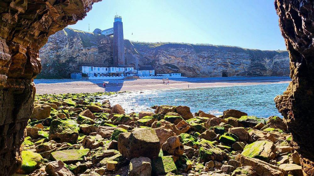 Marsden Grotto in the middle of the beach which is a large white building with a tower stretching to the top of the cliffs. The photo has been taken from what looks like a cave in the side of the cliffs with different size rocks above the sea.