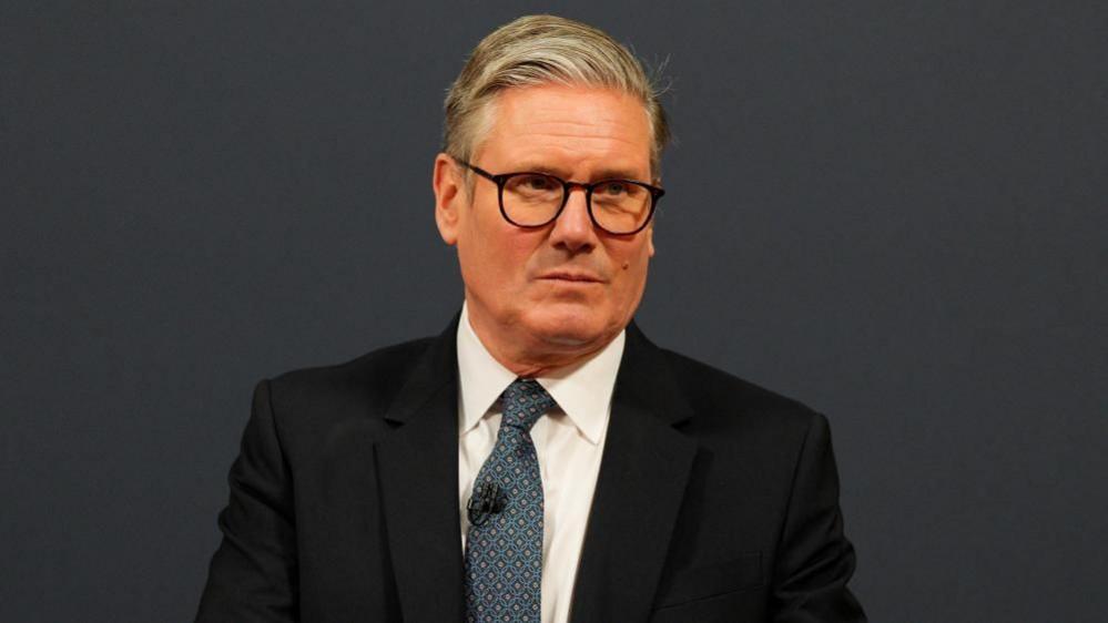 Sir Keir Starmer stands wearing a white shirt, blue tie and black suit jacker in front of a black wall. He has a stern expression. 