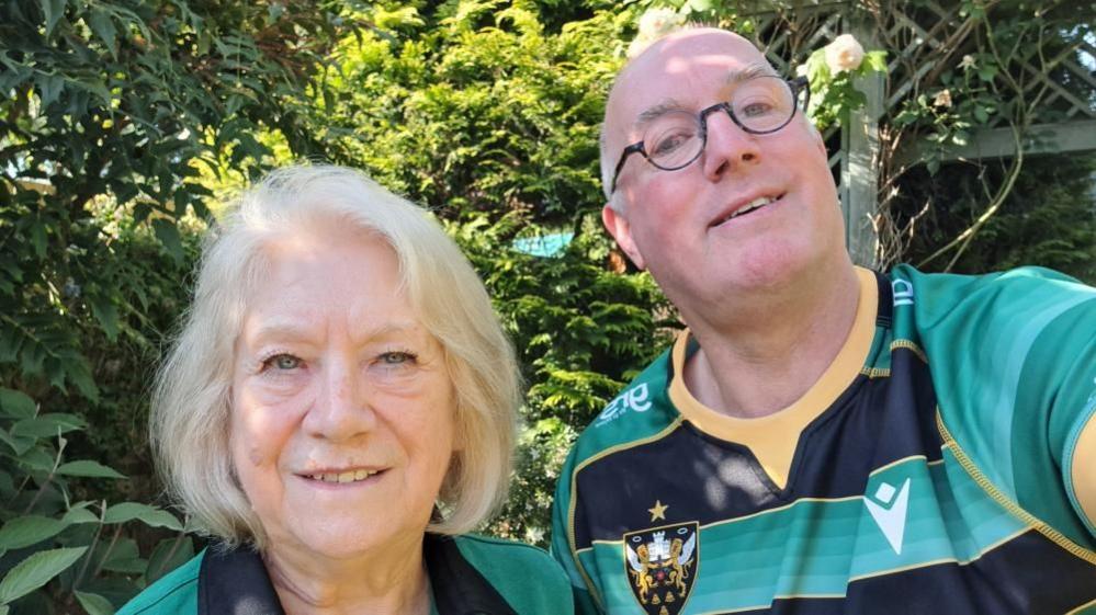 Heather and Chris with Saints shirts in front of a hedge