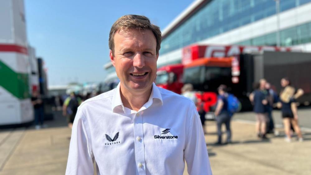 Stuart Pringle in a white shirt, looking at the camera smiling. There are buildings and lorries behind him and people out of focus.