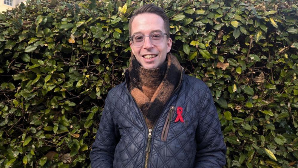 Aled Osborne standing in front of a hedge. He is wearing spectacles, a blue coat and a brown and black scarf.