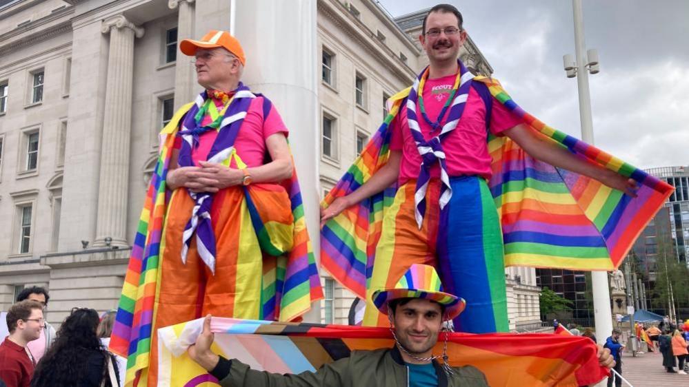 Two people on stilts with Pride flags wrapped around them and trousers made from the flag too. A man in standing in front of them he is wearing a pride hat and holding a flag behind him.