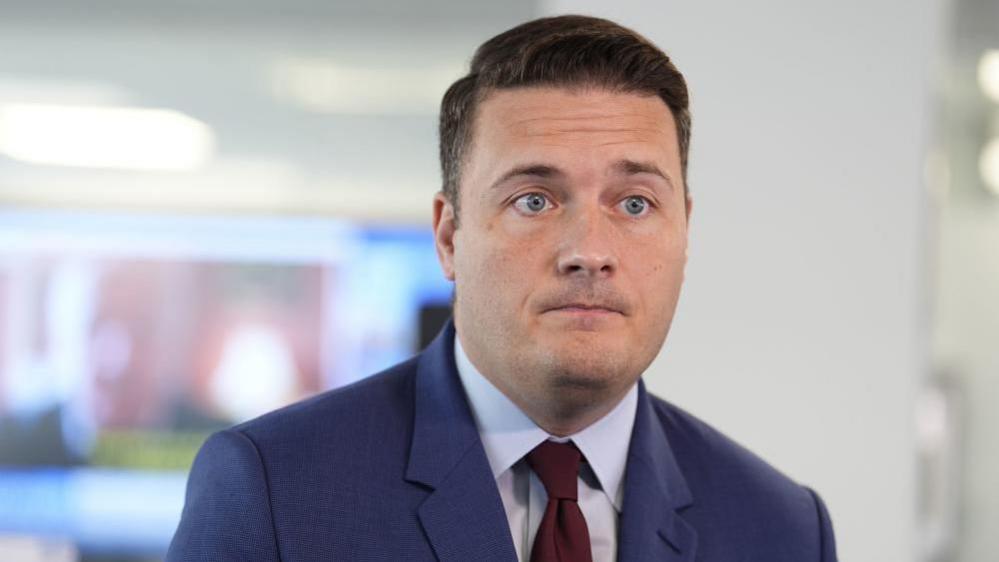 Wes Streeting has short brown hair and is wearing a blue suit jacket with red tie.