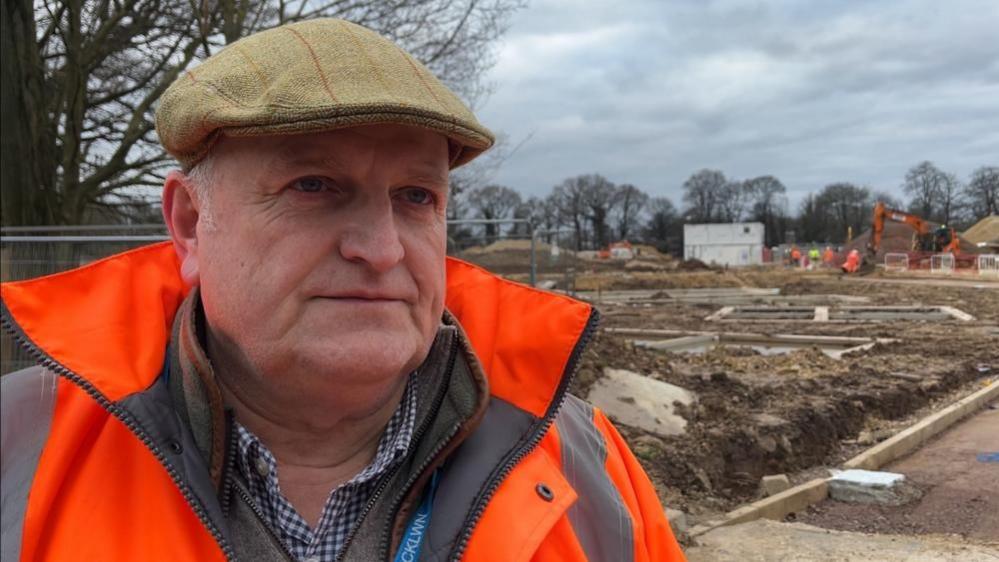 Alistair Beales is standing next to a construction site. He is clean shaven, is wearing a tweed flat cap, and a high viz orange jacket, with a fleece and checked blue and white shirt beneath.