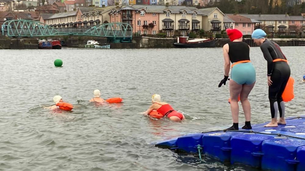 Three people swimming in the harbour, while two people look on from the banks.