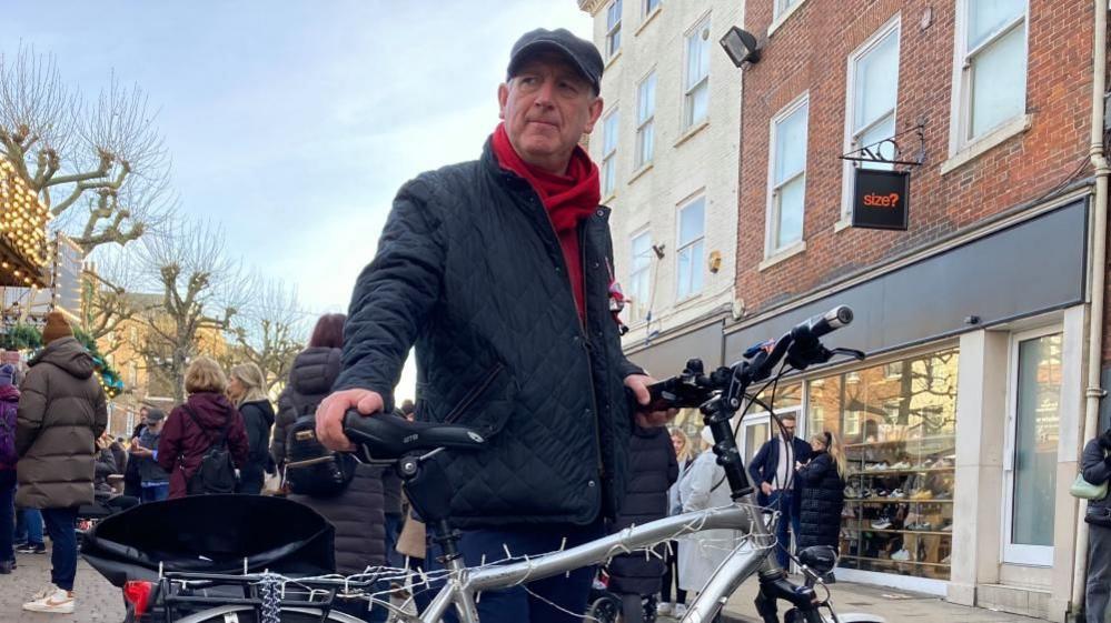 A man holding a silver bike in York city centre. He is wearing a navy puffer jacket and red scarf with a grey hat.