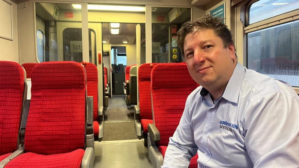 James Steward in a blue shirt, sitting on the red upholstered seats of the Gatwick Express-dressed train that appeared in Nightsleeper