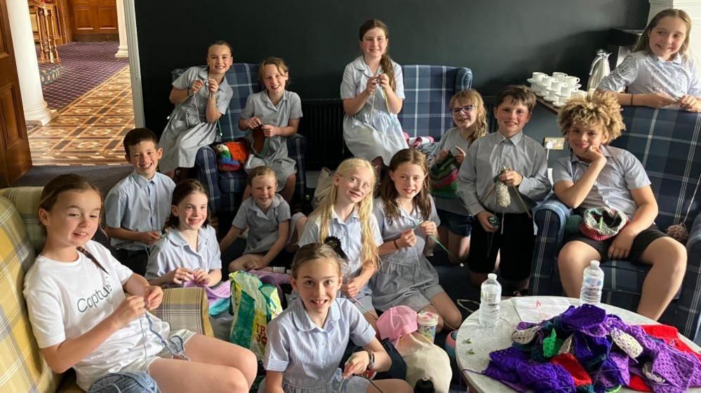 A group of children, all knitting, smiling at the camera.