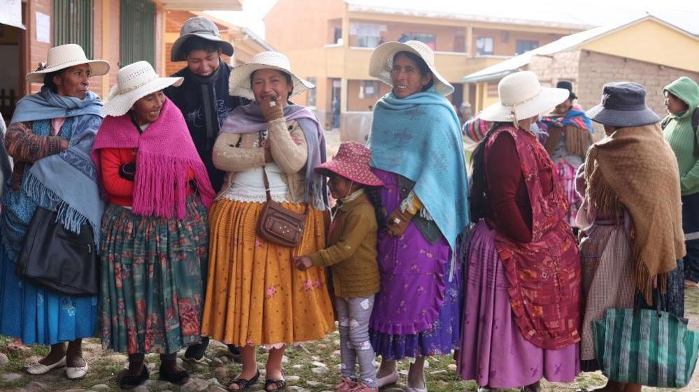 At least seven women in a line, smiling and laughing at the camera, wearing traditional, colourful Bolivian clothing, waiting to cast their vote in La Paz.