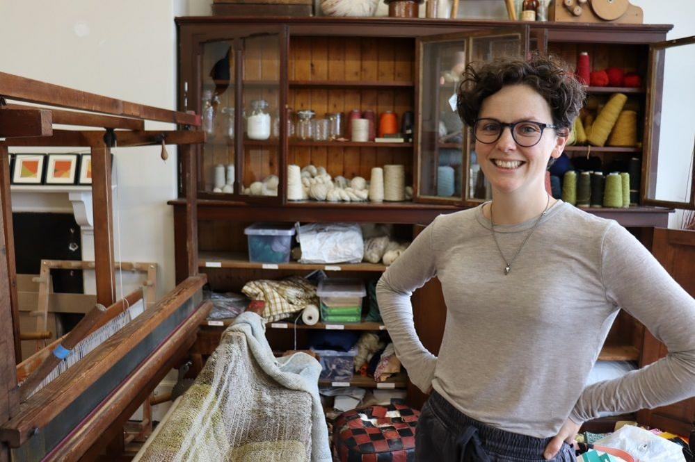 Emily McIlwaine smiling in a grey tshirt beside a large wooden loom with textiles across it
Lots of yarn and threads pictured on shelves in the background