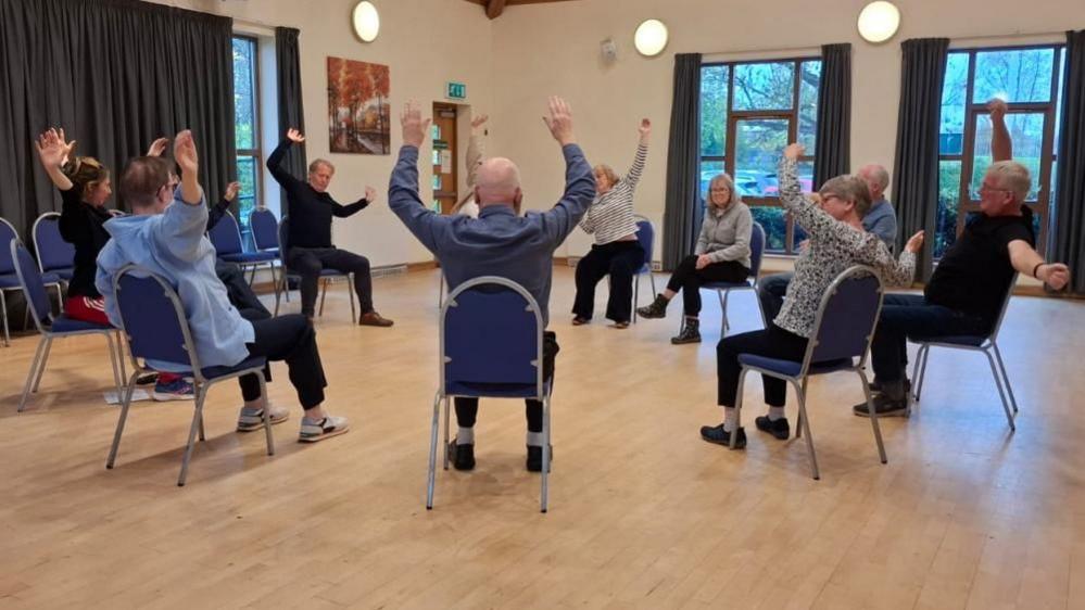 A group of people sitting on chairs in a circle with their arms in the air, taking part in a dance session.
