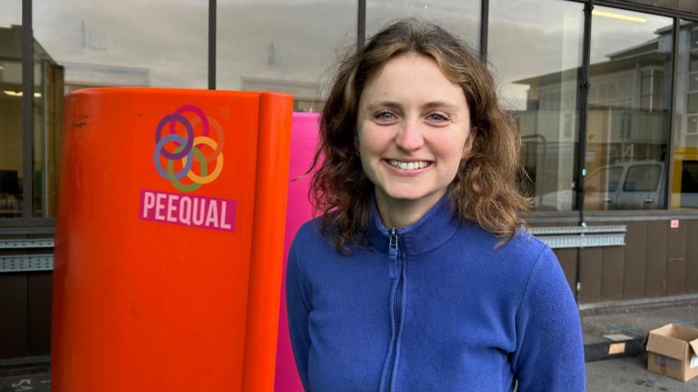 Hazel McShane from PEEQUAL, who is wearing a blue zip fleece, and has shoulder-length wavy brown hair. She is standing in front of an orange panel, which is the side of one of the women's urinals, and displays the logo of the company.