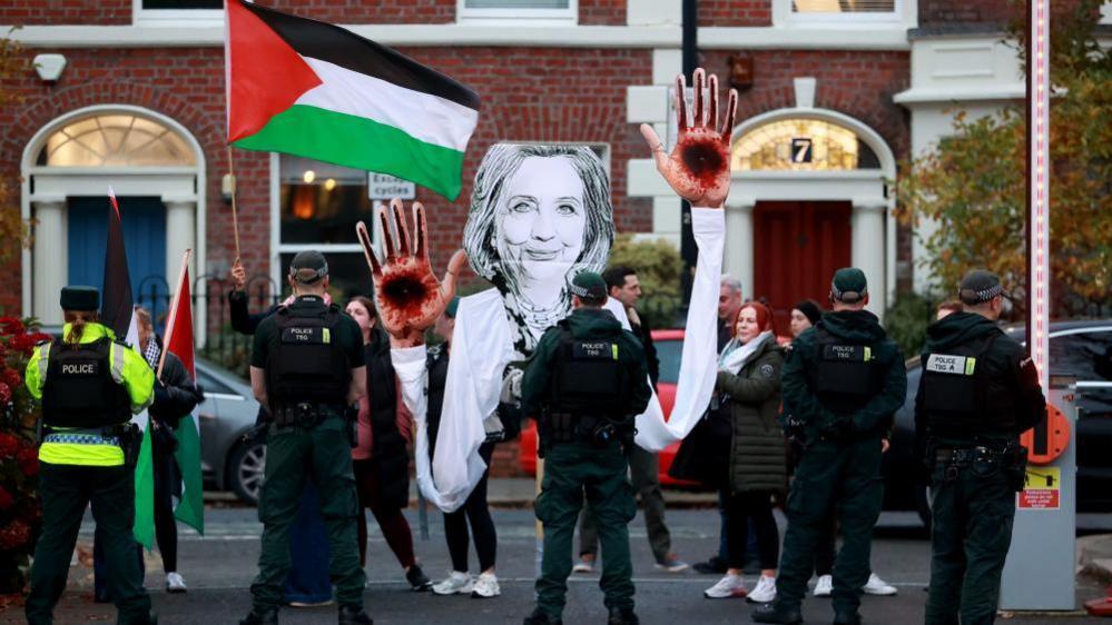 Protestors are pictured standing behind police holding a Palestinian flag and a large black and white cut out of Hilary Clinton with blood on her hands.