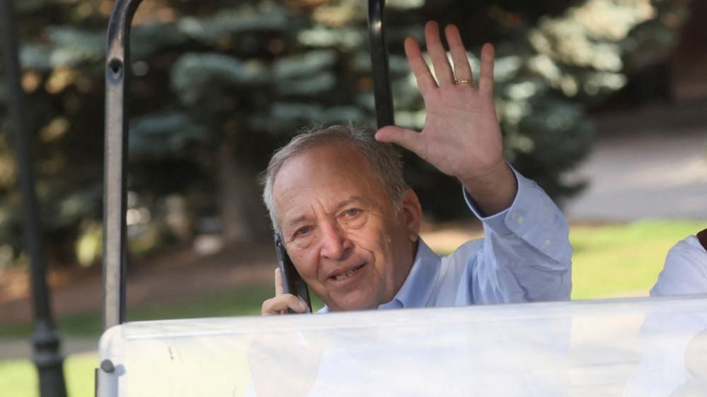 Former Harvard president Larry Summes waves from the seat of a golf cart as he talks on a mobile phone
