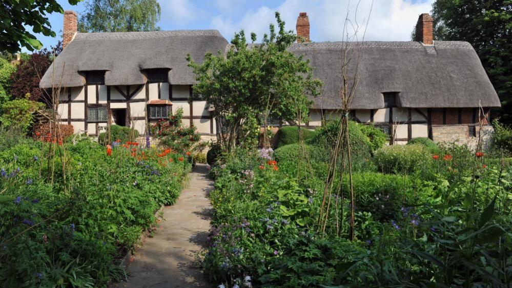 The childhood home of Anne Hathaway, a picturesque thatched cottage, sits in the background behind a garden on a bright summer's day. 