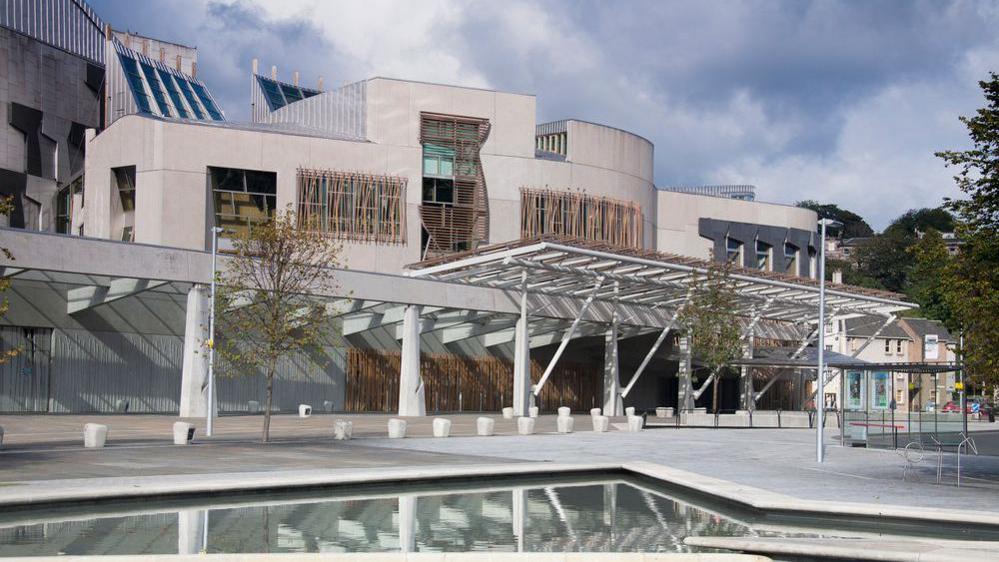 The exterior of the Scottish parliament building in Edinburgh. The building is a beige/grey colour with a walkway in front of it as well as a water feature which reflects the building.