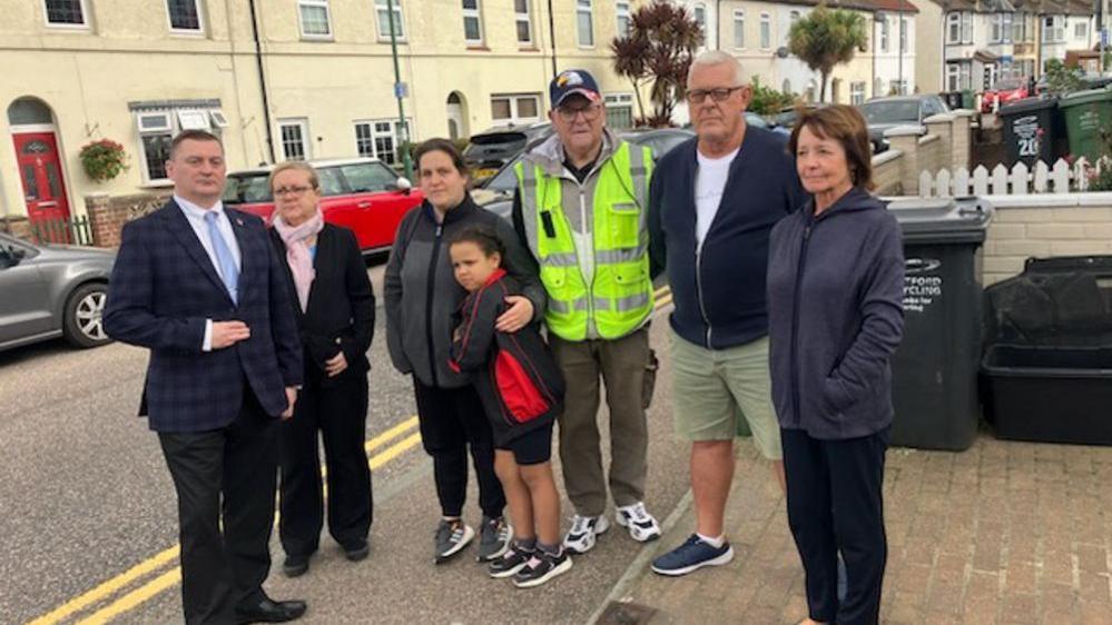 A man in a business suit, a woman in a dark suit and pink scarf, a mother and young son in dark clothing with their arms around each other, a man wearing a hi-vis jacket, a man in a white t-shirt, blue jacket and tan shorts and woman in dark clothing stand side by side in Stanhope Road, a suburban street of two up, two down houses in Swanscombe.