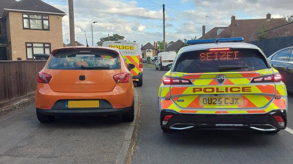 An orange car is stopped on a pavement of a residential street. A police car is next to it on the road with the words 'SEIZED' on a digital display. Behind the orange car is a police van.