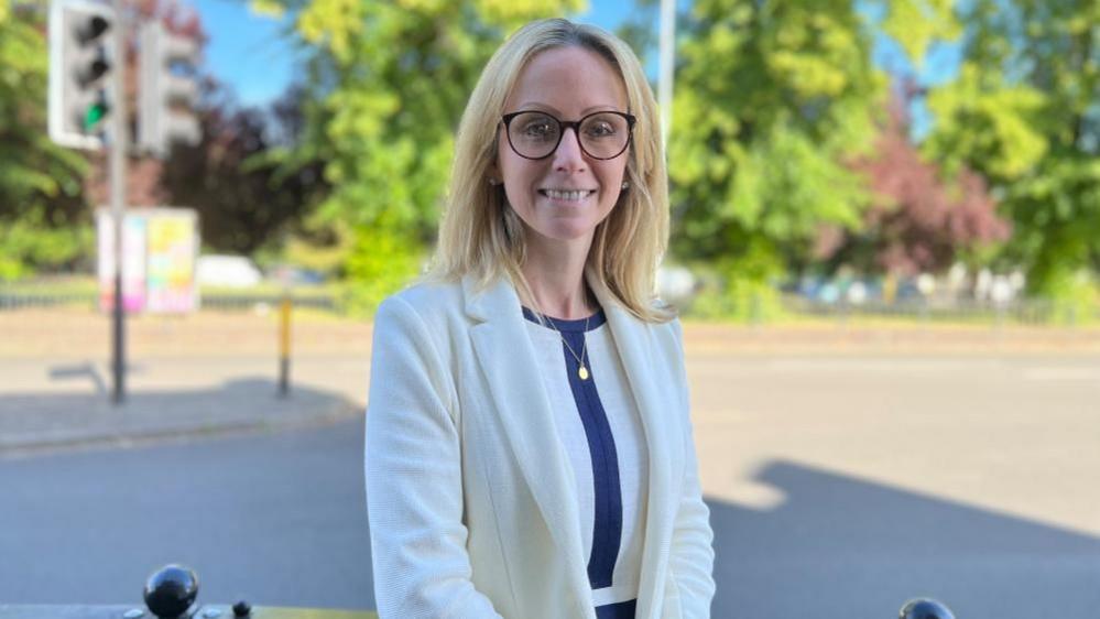 Lucy Rigby with long blond hair and glasses, smiling at the camera and wearing a white jacket and white top with blue stripe down the centre. A road with traffic lights and trees is visible behind.