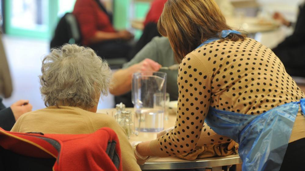 A pensioner wearing a pale yellow jumper is having a meal while seated at a table, facing away from the camera, with a woman next to her wearing an apron and either putting something on the table or moving it away. A blurred partial image of someone eating can also be seen at the other side of the table, along with several people in the background