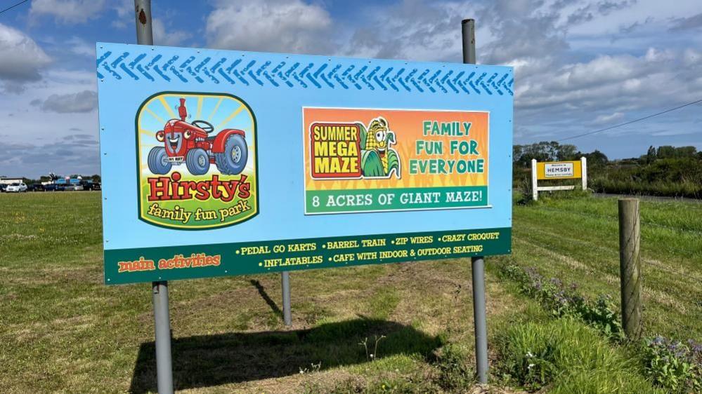 A blue sign in a field reading "Hirsty's Family Fun Park" with a graphic of a red tractor