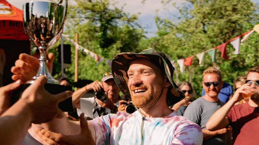 Tom Wheeler - with a ginger beard and wearing a camouflage, floppy hat and tie-dyed T-shirt - claiming his trophy