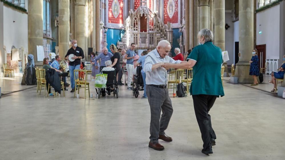 A woman in a nurse's uniform and wearing a face mask dances with an old man. In the background people are sat on chairs playing instruments like the tambourine. They are in a grand-looking monastery.