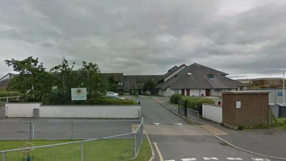 A Google streetview of Ysgol Friars school. It is a cloudy, grey day and you can see the school buildings in the image. Along with a school sign in the green shrubbery to the left of the image. There is also a road which leads to the car park.