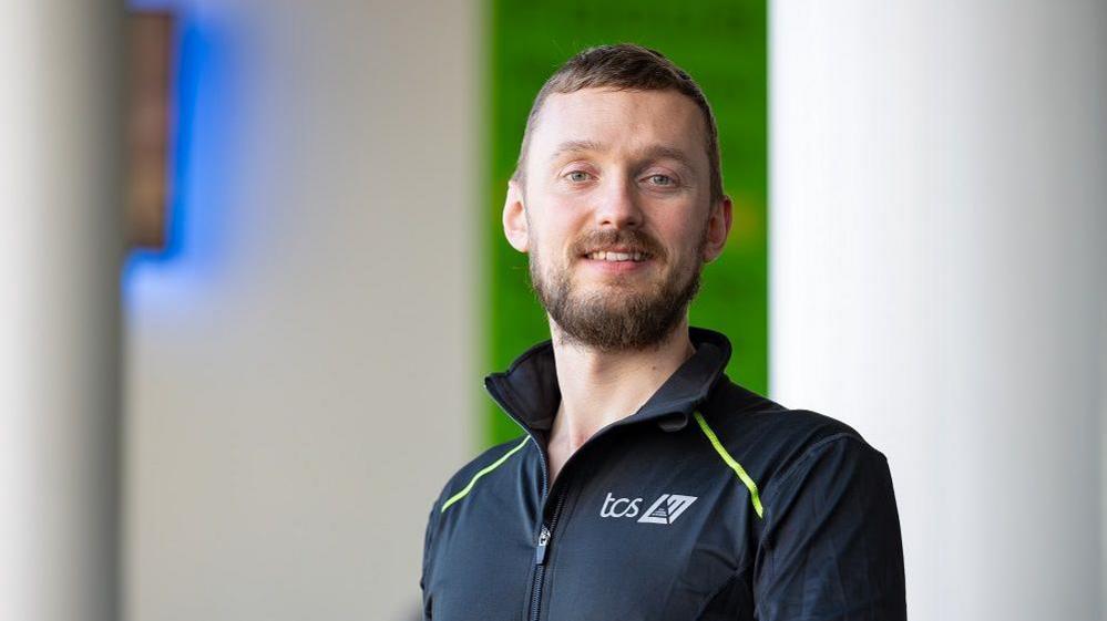 Mark Learmouth smiling at the camera. He has short brown hair and a brown beard and is wearing a sports top.