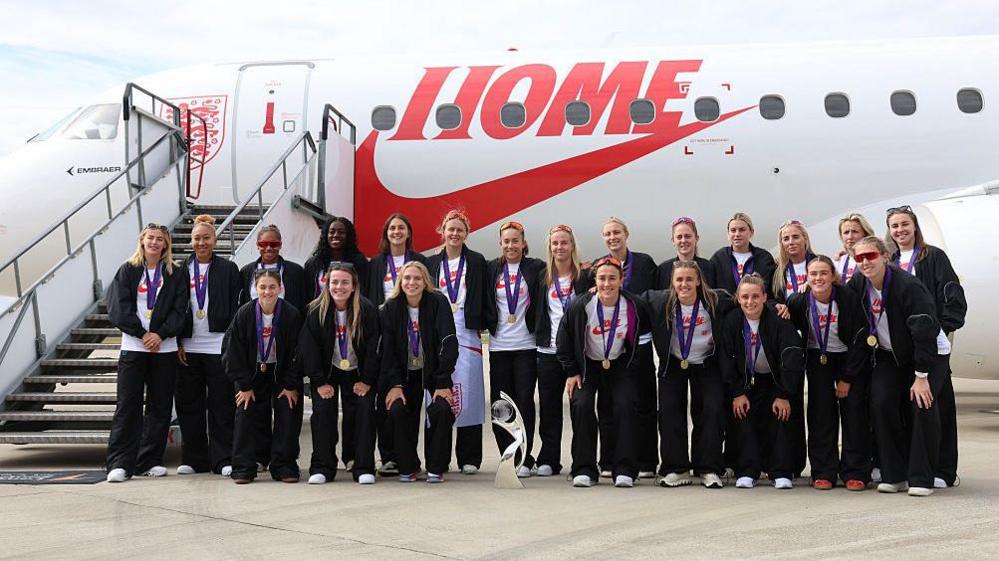 England women's team pose in front of their plane with their winner medals around their necks