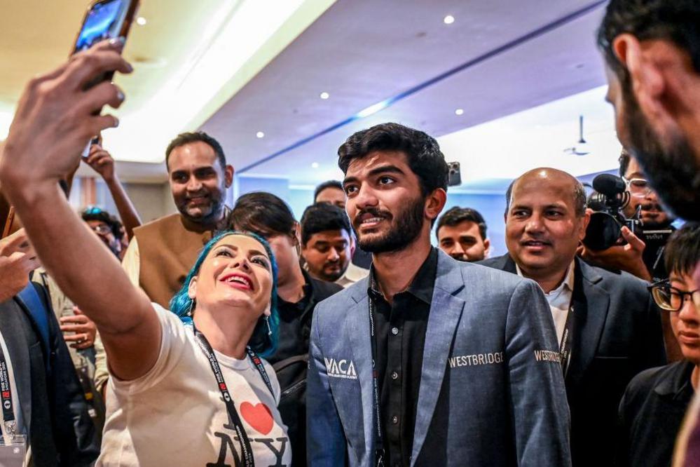 Gukesh poses for a selfie with chess fans, he is wearing a blue blazer and black shirt. He is stood next to a woman and is surrounded by people behind.