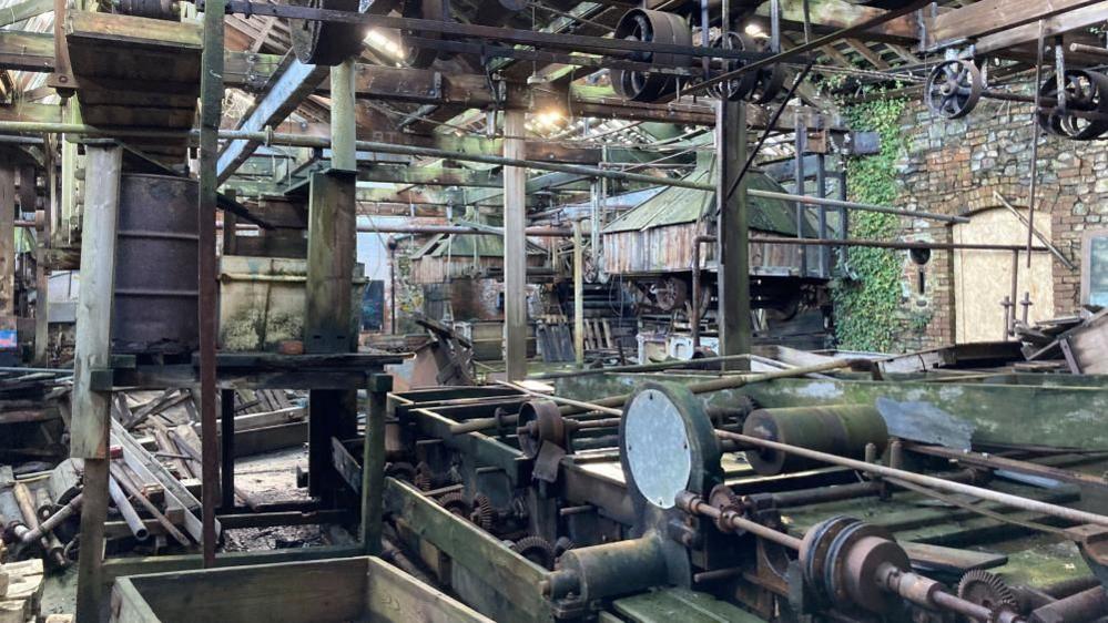 The interior of the 18th century Tone Mill in Wellington, Somerset. Exposed brick walls are overgrown with Ivy and support dilapidated wooden machinery. The floor is strewn with debris and building materials. Rusted and broken machinery fills all corners of the site. 