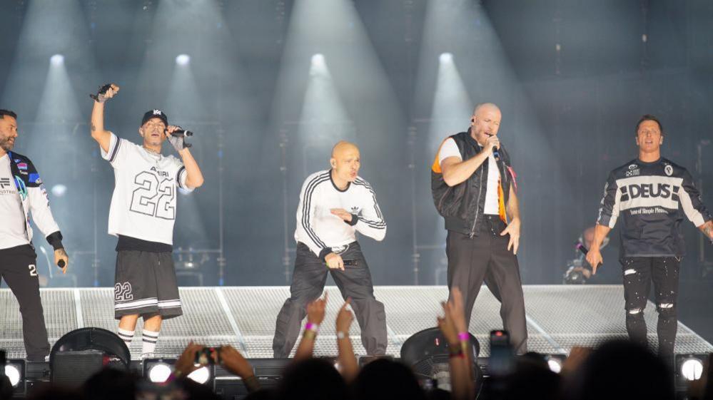Five members of the boy band are under bright white lighted stage. Each of the members are holding a microphone and appear to be singing. There are people on the bottom of the image watching and filming.