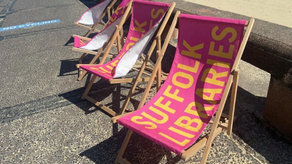 Pink deck chairs are lined up in a row facing the camera on the promenade at Lowestoft. They have big yellow writing on them saying Suffolk Libraries