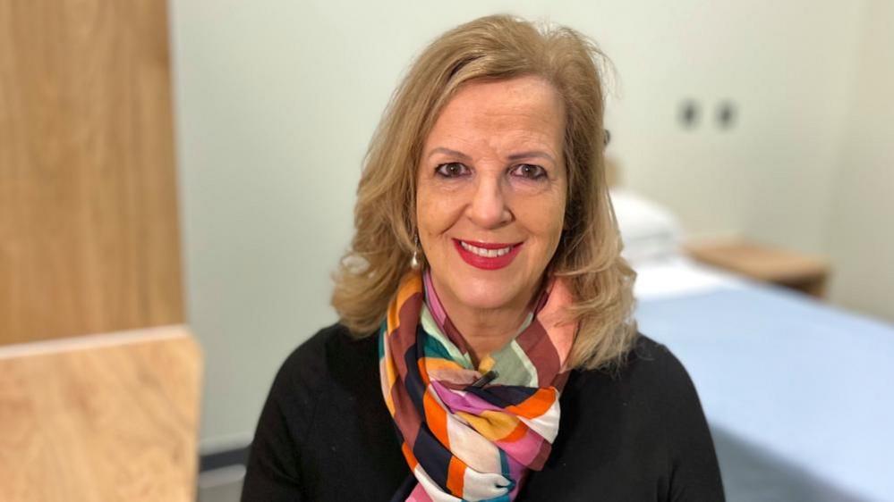 Caroline Donovan is wearing a black jersey and multi-coloured neck scarf. She has shoulder length blonde hair and is wearing an earring. She is standing in a hospital ward, with a bed and other furniture around her.