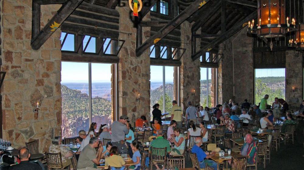 Dining room with high ceilings and windows overlooking Grand Canyon, where people sit to eat or stand, during the day