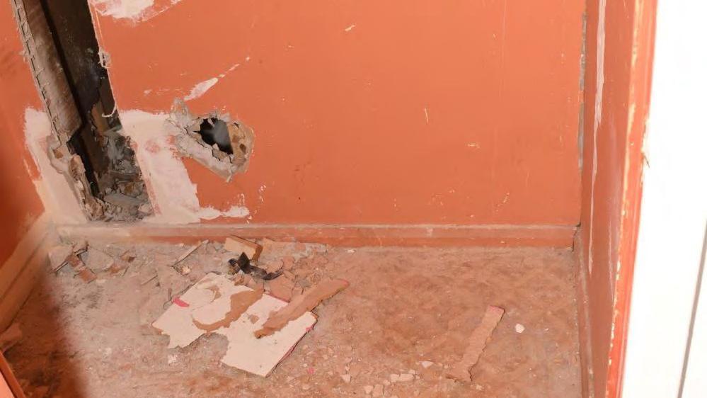 An orange wall with hole and gaps and debris on the floor. The space behind the wall can be seen