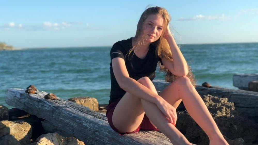 A young women with long fair hair sits on a log in front of the ocean. 