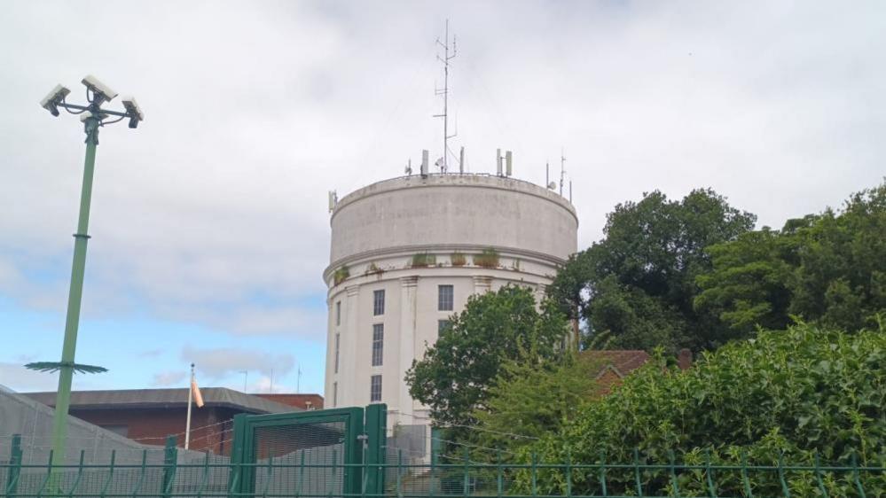 A round, white water tower set in grassland but surrounded by other water works buildings, trees and green metal security fencing.